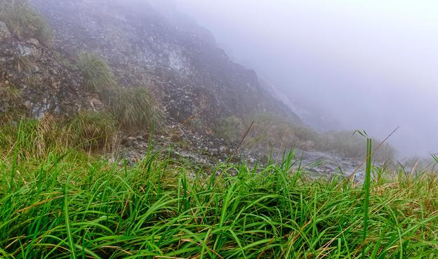 Volcanic gases from a fumarole in a mountainous landscape with grass in the foreground.