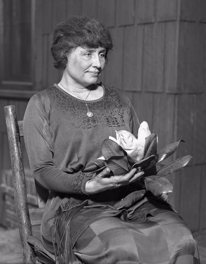 Photograph of Helen Keller taken circa 1920. Helen sits in a wooden chair holding a large magnolia flower. She wears a dark dress and a necklace. 