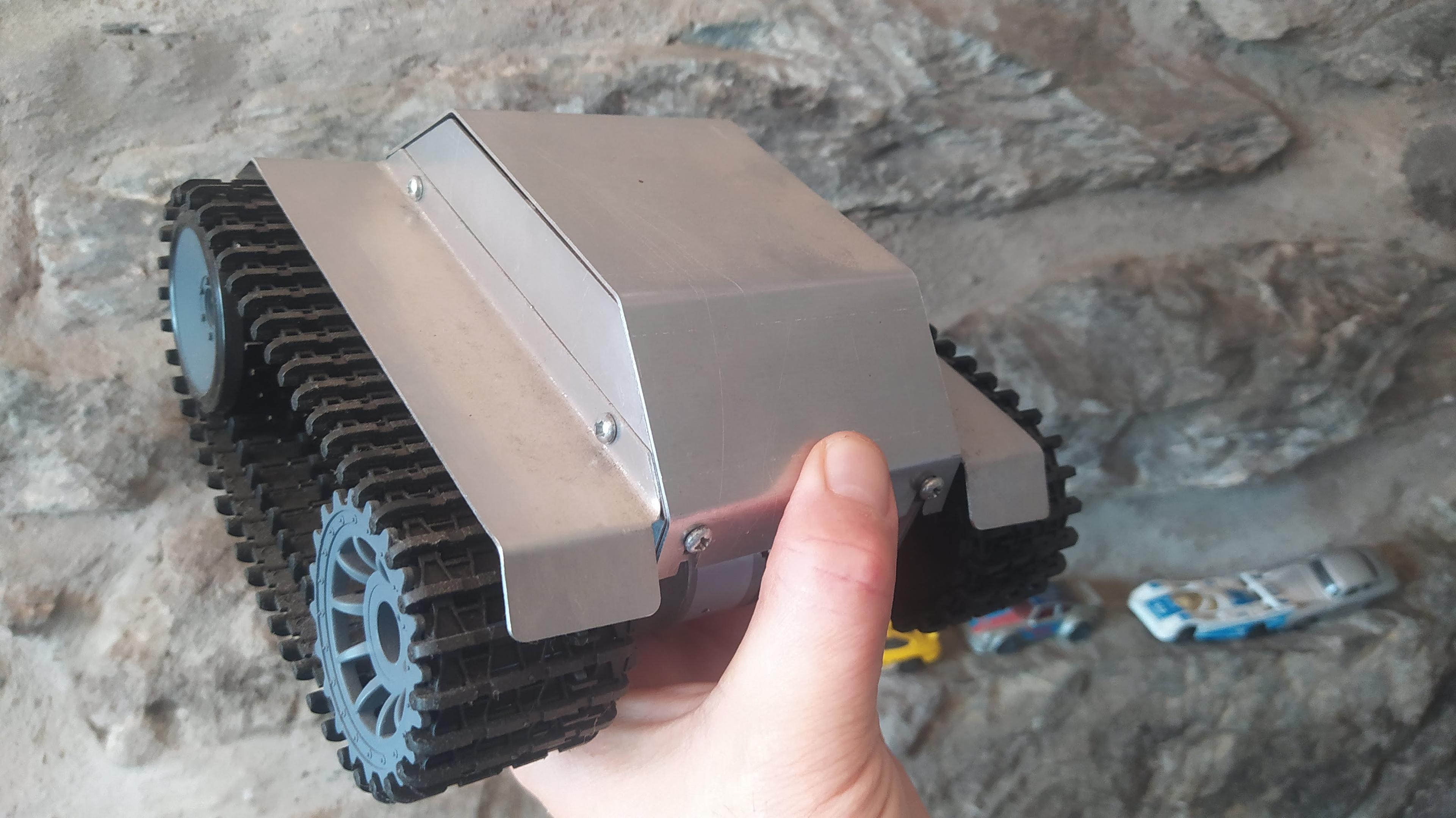 My hand holding a small (circa 20cm square) tracked robot rover. It has an aluminium body shell cut and folded and bolted together. Looks slightly mandolorian! 