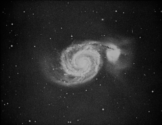 A grainy, black and white image of two galaxies. The larger of the two galaxies, a spiral, is on the left side of the image. Two well-defined arms extend to from its center and form loose spirals around the middle. The smaller galaxy on the far-right side is fuzzy sphere. The view of this smaller galaxy is partially obstructed by the tip of one of the arms of the spiral galaxy, which appears to cover the top third of the smaller galaxy. Many white dots, stars, are seen on the black background.