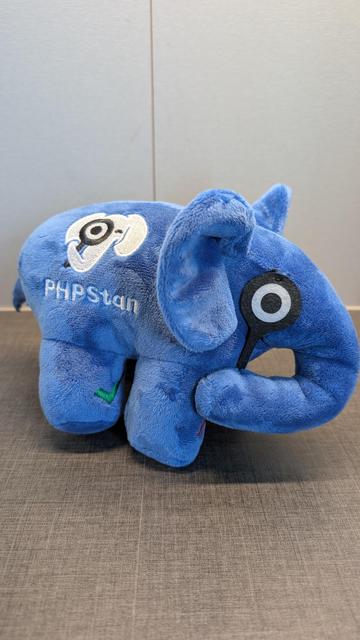A blue plushie elephant with a design inspired by the well-known PHP statical analysis tool "PhpStan". 
It carries a magnifying glass above one of its eyes, held by its trunk. The other eye is closed to a wink.
Two of the four feet show green check marks, while the other two show red cross marks. 
The body shows the tools logo and name on one side and the PHP logo on the other side.