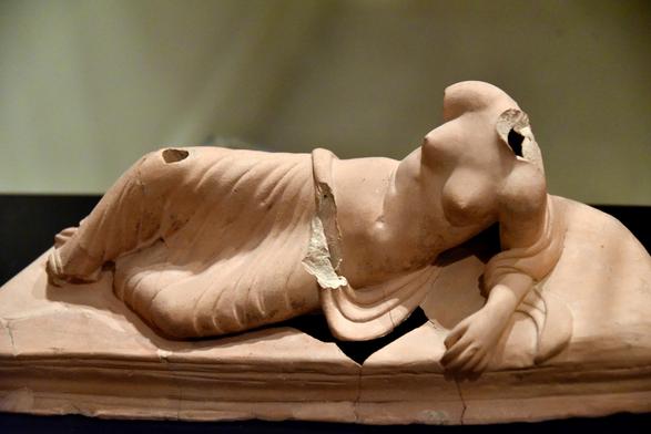 Terracotta figurine of the goddess on a large couch (klinē), leaning on her left elbow. This was the typical position for dining at a banquet (symposium), which was derived from the Greek tradition and was adopted by the Romans. The hole in the upper surface of the right knee possibly was a place to insert a lamp.