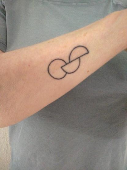 Zu sehen ist ein Tatoo auf dem Unterarm. Es zeigt einen geschlossenen Kreis und zwei versetzte Halbkreise, von welchen einer in den geschlossenen Kreis hineinragt. Sie bilden eine Kette. Im Hintergrund ist ein graues T-Shirt zu sehen. 