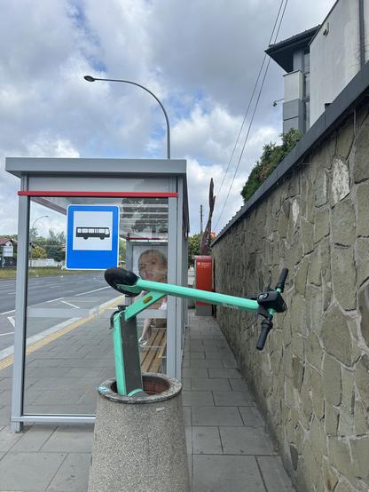 There is a bus stop in the backdrop of the picture. Front and center is rubbish bin with an electric scooter jacked vertically inside it and standing out. 
