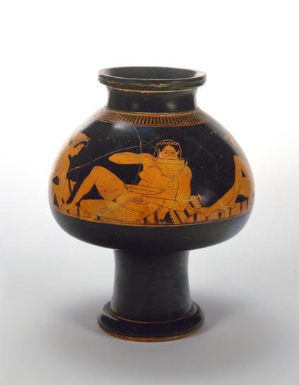 Red-figure vase painting on a psykter, a vessel that was filled with wine and then set inside a crater holding cold water to keep it cool. In the artwork decorating the psykter, four naked women are lying on a couch with richly coloured cushions and mattresses. We only see one fully in this photo, Palaisto, whose face looks directly out at us, drinks from her cup. Inscriptions provide the names of the women and the craftsman who created the vase, Euphronius. We see a set of legs on the right an…