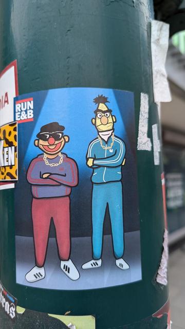 Aufkleber an Ampelmast: Ernie und Bert gezeichnet, gekleidet wie Run D.M.C in Trainingsanzug, mir Goldkette, Ernie trägt Sonnenbrille und schwarzen Hut, Bert schaut skeptisch, beide verschränkte Arme. Oben links Logo: "RUN E&B".