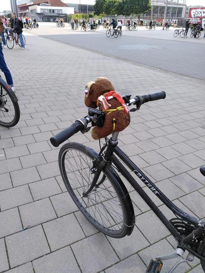 Plüsch-Elch Gunvald auf dem Fahrradlenker. Im Hintergrund Menschen auf Fahrrädern und das Millerntor-Stadion 