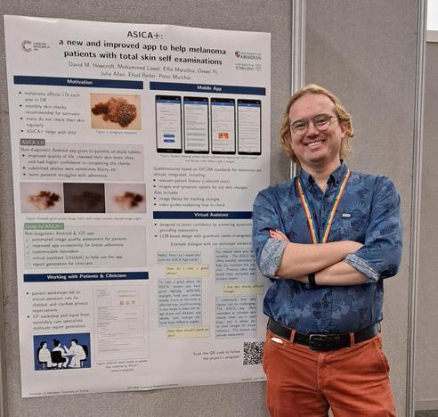 a blond haired person in a blue shirt and orange trousers poses next to their academic research poster