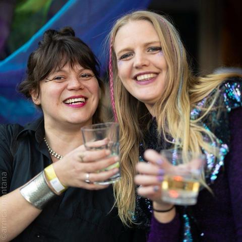 Two women are smiling and holding possibly nonalcoholic drinks, posing together in a colorful setting with vibrant decorations in the background. One woman has dark hair and bold makeup, while the other has long, light hair with colorful highlights.