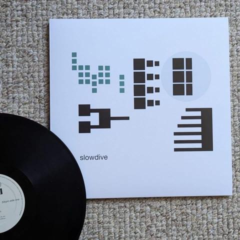 Eine schwarze Schallplatte ist teilweise neben einem weißen Albumcover zu sehen, das geometrische Formen und das Wort "slowdive" zeigt. Der Hintergrund besteht aus einer strukturierten beigen Oberfläche.