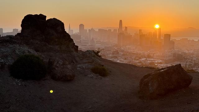 Barren rocks in the foreground frame a city skyline. The sun rises above some towers illuminating the haze. 