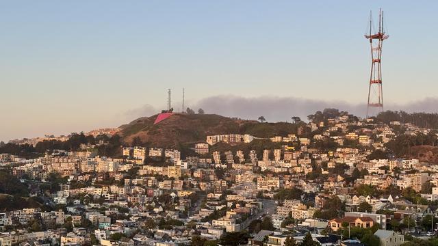 Numerous buildings wrap a mountainous ridge. A green prominence features a large pink triangle, another supports a large red radio tower. 