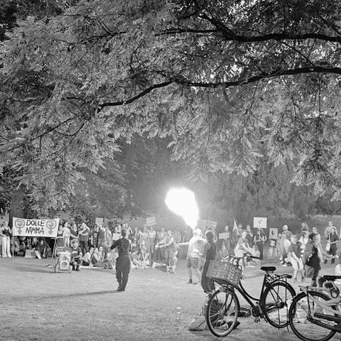 Een groep mensen is bijeen in het Kronenburgerpark tijdens een demonstratie tegen straatintimidatie. Op de voorgrond staan enkele fietsen geparkeerd op het gras. In het midden van de afbeelding voert een vuurspuwer een act uit, waarbij een grote vlam uit zijn mond komt. Op de achtergrond houden deelnemers borden omhoog met feministische symbolen en slogans, waaronder één met de tekst "DOLLE NIMMA". De scène speelt zich af onder een grote boom met uitgestrekte takken.