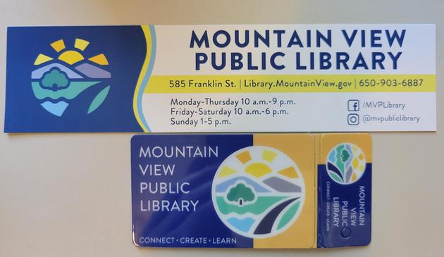 Photo of a Mountain View Public Library card and bookmark. They rest on an off-white desk mat. 

The library card has a background of solid blue (left 70%) and yellow (right). A circular logo overlaps the boundary of the backgrounds, with an illustration of a sun casting rays from partially behind two hills, a green deciduous tree in a green grassy area, a small section of water, and a dark-blue road on the right. Text at left reads "Mountain View Public Library" in large white block letters. Text at bottom reads "Connect Create Learn" in white block letters. Atrached to the library card, at right, is a small keychain card with identical elements but laid-out so the circular logo is on the left and text at right.

The bookmark has a blue (leftmost 25%) and white solid background (right), divided by a gently-curving border of blue and yellow lines. The circular logo described previously is on the blue background, but the border lines around the elements is blue instead of white. Text on right reads, "Mountain View Public Library" (large blue block letters). Text that follows lists the address, hours, and social media addresses.