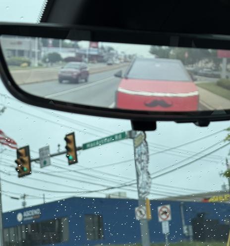 A rearview mirror captures a view of a Tesler Cybertruck with red wrap and a large mustache decal on the front end