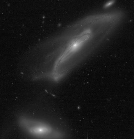 A greyscale image of two interacting spiral galaxies. The larger galaxy in the upper part of the frame has a bright central core surrounded by a long bar structure. The bar is crossed by dark dust lanes. Emerging from the bar is the beginning of one spiral arm at about 6 o'clock that curves upward. There are additional, barely visible spiral arms that wrap around the galaxy. The smaller galaxy in the bottom left portion of the frame also has a bright core, surrounded by an oval-shaped central region, and very faint, wispy spiral arms. The black background of space is filled with foreground stars and tiny background galaxies. The image has a noticeable amount of detector noise which has not been removed. 