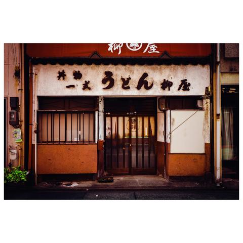 This color photo shows the exterior of a well-worn udon restaurant in Nagoya. The façade features a signboard with old-style Japanese calligraphy that reads “うどん” (udon), flanked by other characters indicating a set-meal shop. The building’s walls are painted in a warm mix of white and brown, both showing visible wear and patina. A pair of barred windows flank a central entrance with a traditional translucent noren curtain. Above the door is a faded red awning bearing the restaurant's name, partially obscured by time.