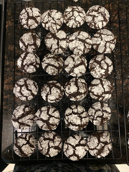 Viewer is looking down on a black wire rack filled with cookies, six rows of four. The rack is sitting on a dark brown granite counter. The cookies are dark chocolate covered with confectioner’s sugar. They have beautiful cracks all over which show pleasingly dark through the white sugar.