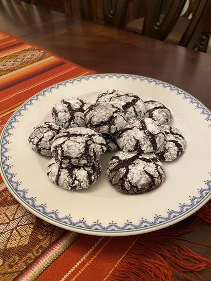 A pile of cookies on a white plate that is rimmed with a delicate blue design bordered by a thin green stripe. The plate sits on runner in shades of red, orange, and burgundy on a dark wood dining table. The cookies are dark chocolate covered with confectioner’s sugar. They have beautiful cracks all over which show pleasingly dark through the white sugar.