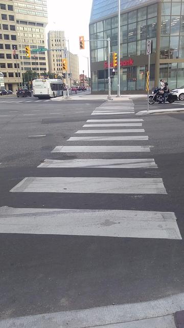 Colour photo of the north-south pedestrian crosswalk on the west side of Main in downtown Winnipeg, looking south, on the first official day the pedestrian access was reopened since 1979-02-24. The white crossing pictogram had just illuminated prior to my first time going through one of the crosswalks: a total of eight lanes need to be traversed along this portion. There are four buildings visible to the south and east, including the art deco-inspired Victory Building, furthest away.