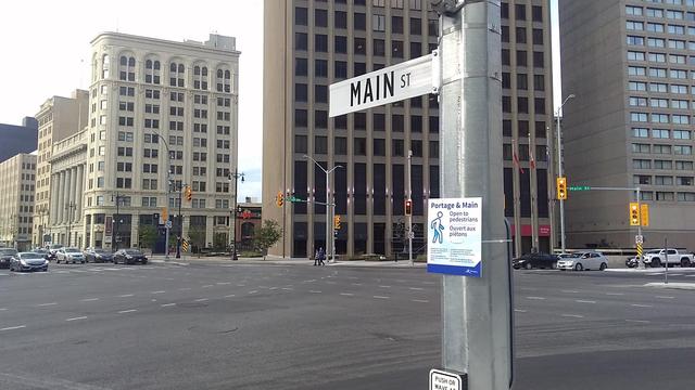 Colour photo looking northeast from the southwest corner of Winnipeg's Portage & Main intersection, on it's first official day of being reopened to pedestrian traffic. A black letter-on- white sign that says MAIN ST is located at a relative eye level on a fresh silver galvanized light pole whilst we see the lower few floors of the Richardson Building and the lower western portion of the Fairmont Hotel. There is a small temporary sign also on the light pole that says:
Portage & Main
Open to pedestrians
Ouvert aux piétons
(omitting small print which just confirms that it is now permitted to cross the intersection as of this date, in both English and French).
