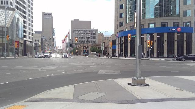 Colour photo looking straight down Portage Avenue in downtown Winnipeg from a seated position on the northeast corner of Portage & Main as several vehicles wait to proceed east or north through the intersection. This spot marks "Mile 0" of the Yellowhead Highway, as is noted by a sign on the right side of the westbound lanes.
There are many flags of Canada, Manitoba, and the City of Winnipeg on flag poles between the west and east lanes of Portage, along with numerous buildings along the roadway, including the Paris Building and Radisson Hotel.