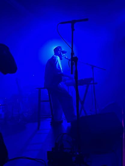 River Tiber is seated at a keyboard, singing into a microphone, surrounded by blue lighting. The scene captures the atmosphere of a live music event, with equipment visible in the foreground.