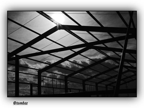 The black-and-white photo shows the metal skeleton of an old industrial hall—empty, raw frames made of beams forming geometric lines and sharp angles. Part of the structure appears weathered by time, with some elements bent or tilted. In the background, a dramatic sky dominates, densely covered with heavy, torn clouds. Sunlight struggles to pierce through their layers, spreading in streaks between dark zones. The entire scene radiates an atmosphere of abandonment, transience, and the beauty found in decay.
