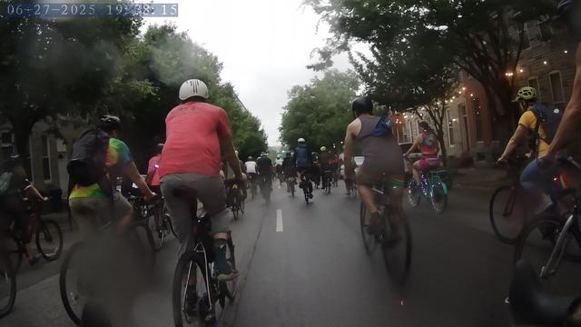A crowd of cyclists, many in rainbow gear, bike through downtown Baltimore in the rain.