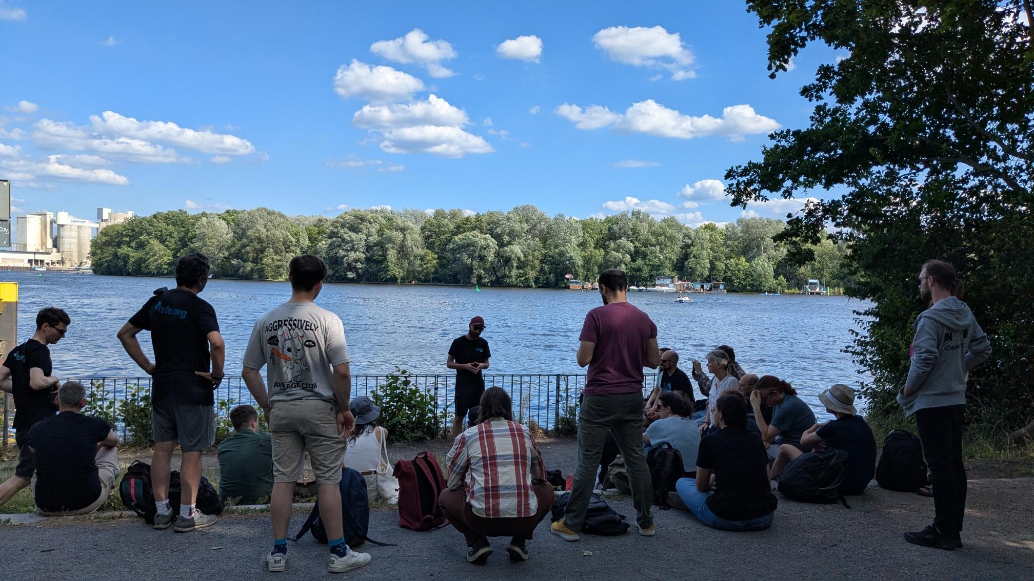 again several people standing in front of one person talking to them. behind water with some boats. on the other side of the water trees.