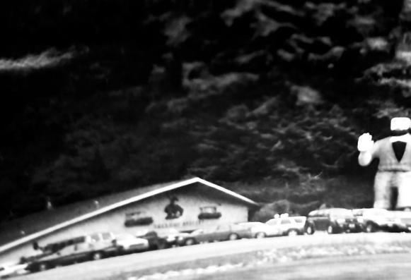 A blurry, overexposed, black & white photo of a “giant” at the edge of parking lot full of cars. There is a low, wide building with an asymmetrical, sloping roof to the left, and a thick woods of tall trees fills the background.