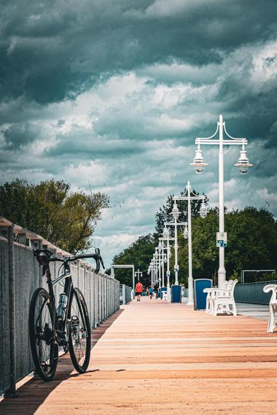 photo of a black Specialized Diverge gravel bike leaning against the railing of a boardwalk. the boardwalk is lined with white lamp posts and white park benches on the opposite side. the sky above is showing dramatic cloud cover 