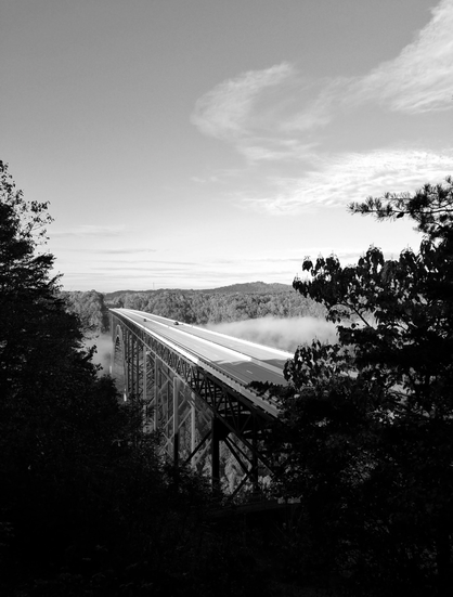 Black and white photo of the New River Gorge Bridge in West Virginia as seen from the boardwalk that allows for scenic views of the bridge and the valley. In the photo there is a layer of clouds just below the bridge.