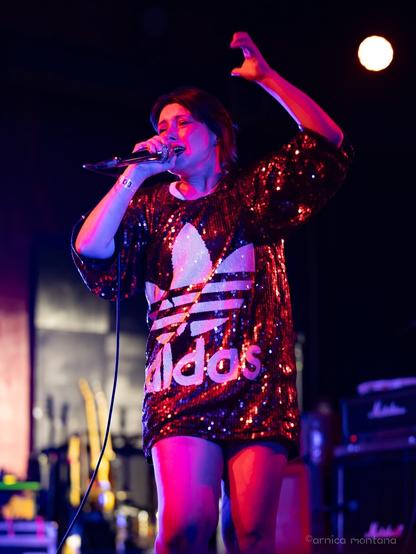 A performer in a sparkly red and black oversized shirt with an Adidas logo is singing into a microphone on stage. Colorful stage lights illuminate the scene, and musical instruments can be seen in the background.