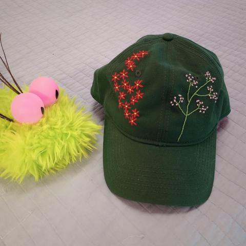 Here's a lovely photo of my green baseball cap. I've embroidered a bunch of red flowers with yellow centers, and another plant with green stems and a heck load of tiny pink blossoms. We can also see Pinkeye. She's a furry green puppet with a pointy pink tongue and pink bulbous eyes. She has two clusters of thin brown feathers popping up behind her eyes.