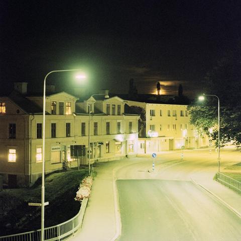 View from a city apartment at night. Street, small apartment blocks and streetlights. The moon can be seen half way above one of the roofs, with a golden glow.