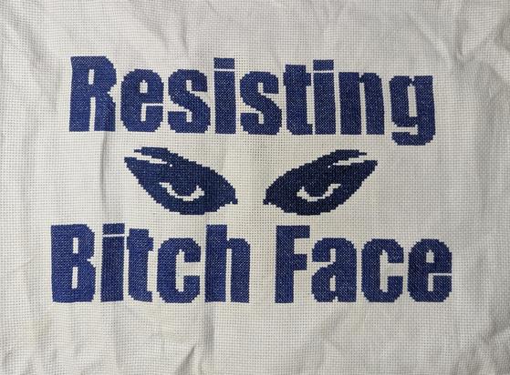 White, wrinkly Aida fabric with dark blue block lettering reading, "Resisting Bitch Face" with the shape of angry eyes in sparkly, dark blue thread.