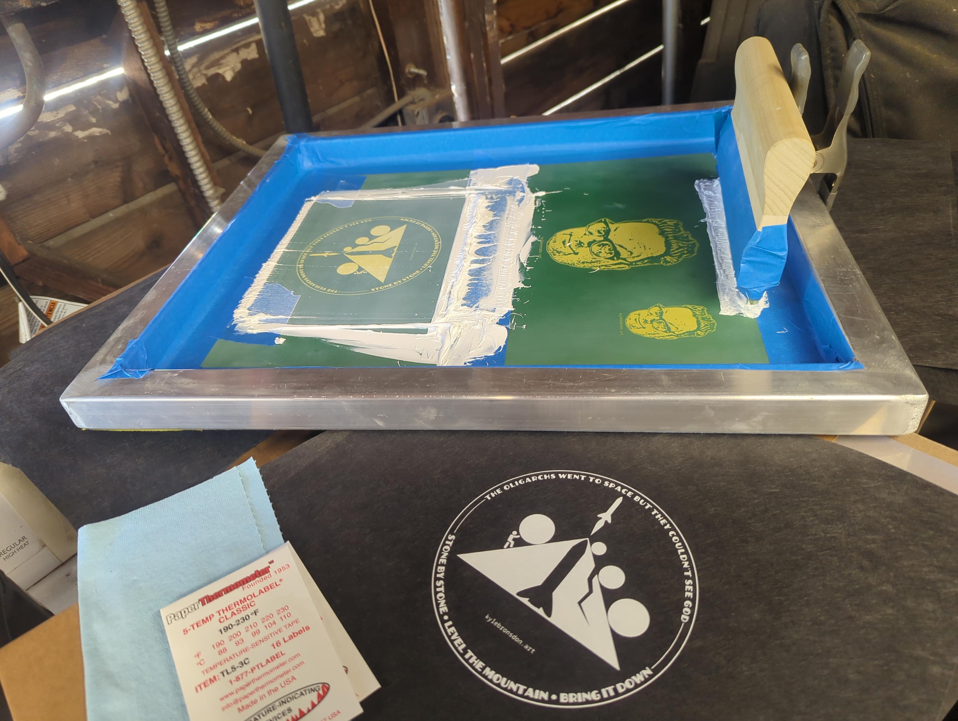A printing screen taped up with blue painter's tape with white ink over the stencil and a taped-up squeegee on it next to some thermal tape and a black pellon printed with the circular design.