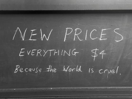 A chalk drawn sign on a café wall that reads "NEW PRICES. EVERYTHING $4. Because the World is cruel"