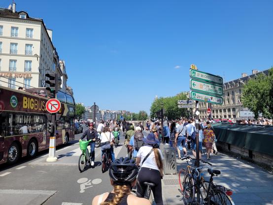 Crowd of cyclists between a tourist double-decker bus and a sidewalk, near Notre-Dame. The sky is cloudless blue.