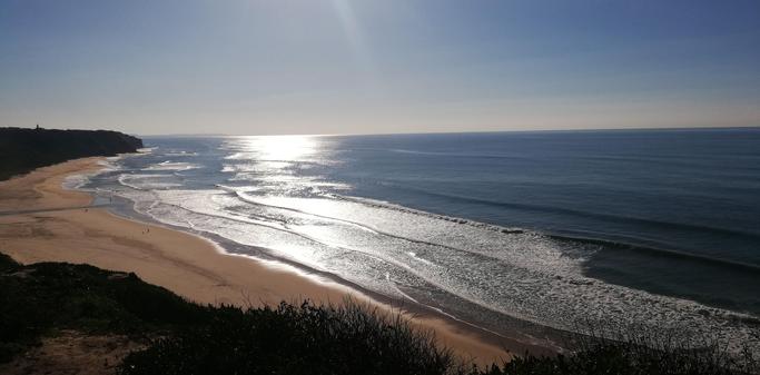 Blick auf eine weite Sandstrandbucht und den relativ ruhigen Altlantik. Die Sonne reflektiert auf dem Wasser und der Himmel ist blau.