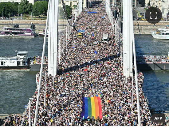 Foto van de brug in Hongarije, van bovenaf genomen. Je ziet een enorme mensenmassa en een grote regenboogvlag in het midden.