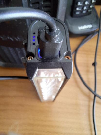 Foto macro de lanterna de bicicleta sendo carregada por porta USB tipo C, apoiada sobre um móvel, próxima a um estabilizador elétrico e a um telefone sem fio. Dá para ver dois parafusos na lateral da lanterna, próximos à porta USB do carregamento, e ambos estão enferrugados