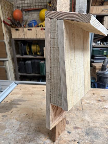 A detailed view of wooden boards secured at a right angle, showcasing their edges and layers. The background includes shelves with various tools and equipment.