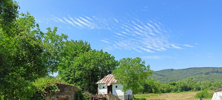 A photograph taken outside on a sunny afternoon. A small building is at the bottom, approximately in the centre. Around it and to the left of it bright green foliage is visible. To the right there is a field and further away some hills. The sky is light blue and there are narrow clouds that look like slightly curved diagonal lines, like something Bob Ross would paint. They are smudged a bit in the lower part of the formation. 