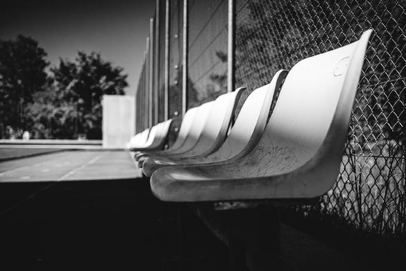 Black and white photo of empty plastic stadium seats lined up against a chain-link fence, with strong shadows and sunlight creating dramatic contrast. The scene appears quiet and still, likely beside a sports court or field.


