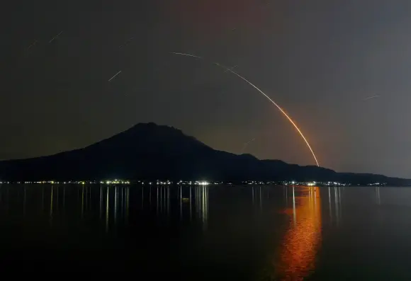 A long-exposure photo of an H-2A rocket at night, the red glow reflected in the water.