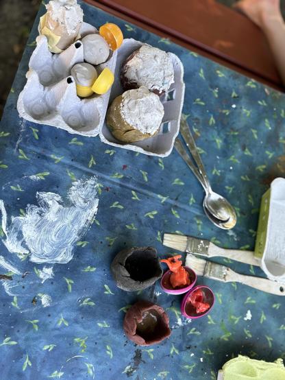 Eier Karton daneben ein paar gebastelte Dinoeier. Daneben Pinsel, ein Löfel und zwei Knetsinoteilformeb. Zwei rote kleine Dinofiguren auch no zu sehen