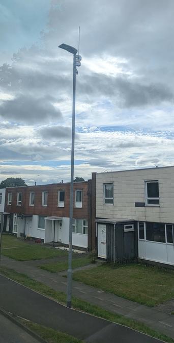 Metal LED street-light pole with a radio antenna, equipment box, and mains power connection in a residential street.