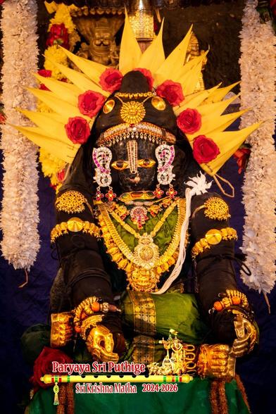 Idol of Hindu deity Krishna in green seated in a yoga position, both legs folded in fully at the knee with the left leg over the right; hands in front over the left leg, palms facing inward. Krishna sits on a platform covered by a darker green silk cloth. He wears gold shoes, gold palm coverings, and gold jewels. A radial arrangement of yellow Ketaki leaves (screw pine) form a halo behind his head. Two thick strings of white flowers hang down on either side of Krishna, slightly behind him, coming from over his head down to his elbow.